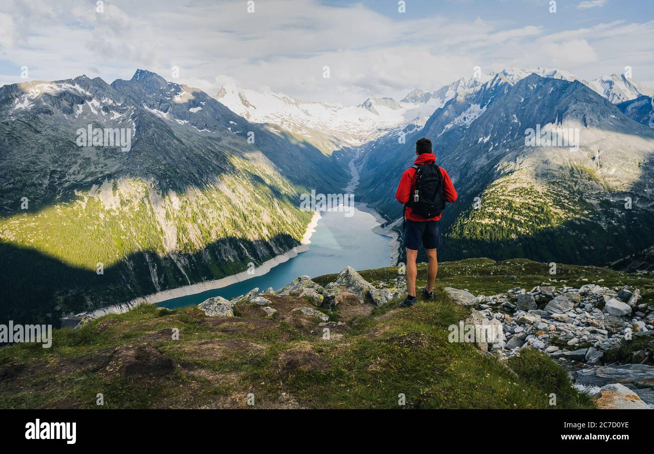 Adventurous man on the edge of a cliff overlooking the beautiful Austria Alps and Lake during a vibrant summer sunset. Taken in Hiking to the Olperer Stock Photo
