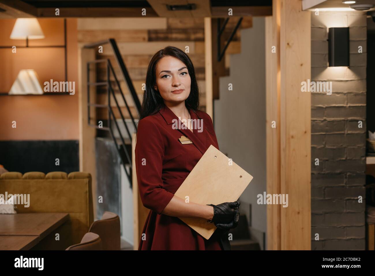 A female restaurant manager in black disposable medical gloves is posing holding a wooden menu in a restaurant. A kind cafe owner in a ruby dress. Stock Photo