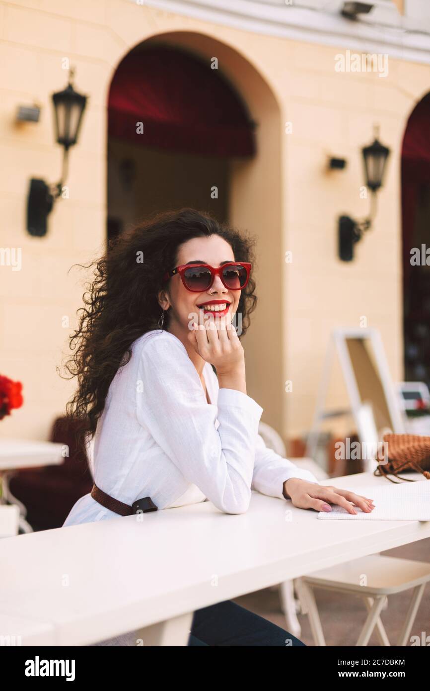 Beautiful smiling lady with dark curly hair in sunglasses standing and happily looking in camera while spending time in summer terrace of cafe Stock Photo