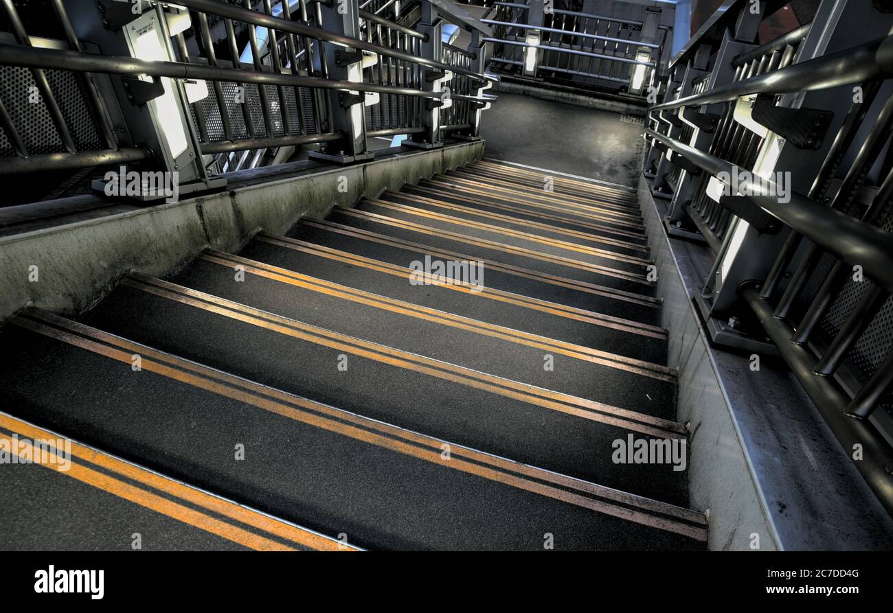 Looking down a dark concrete staircase with two bright yellow caution strips on the edge of each stair and metal railings Stock Photo