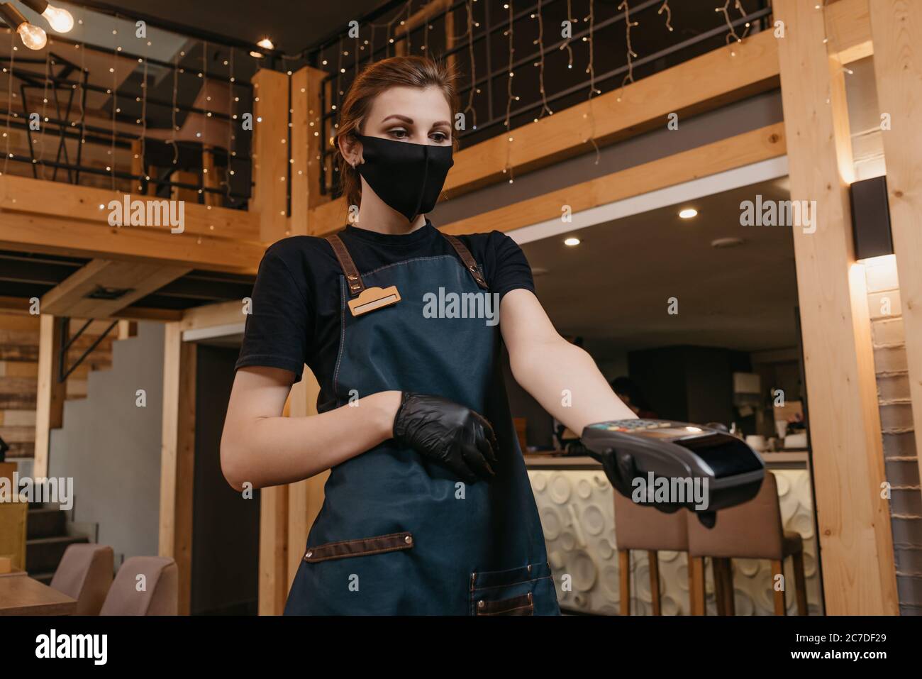 A waitress who wears an apron, a medical face mask, and disposable medical gloves is handing a wireless payment terminal to a client in a restaurant. Stock Photo