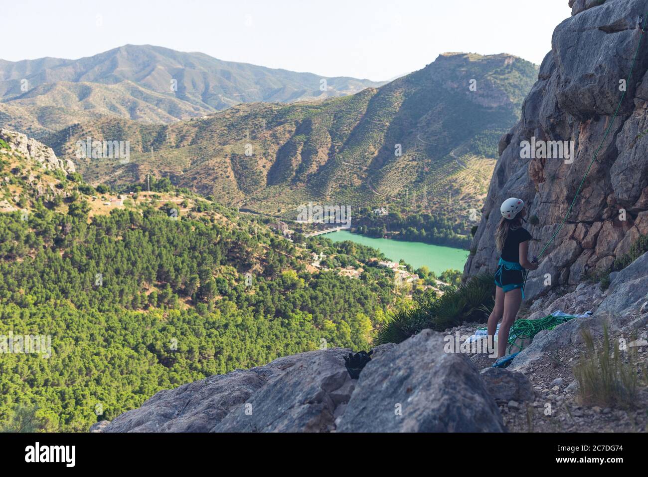 Wide shot of a female climbing a rocky hill surrounded by green mountains and a beautiful lake Stock Photo