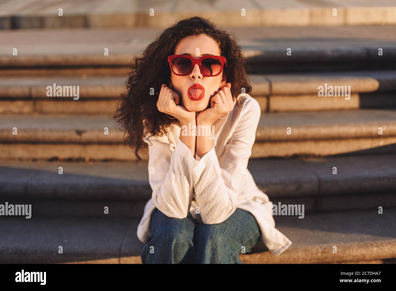 Young pretty woman with dark curly hair in sunglasses and white jacket sitting on stairs on street and dreamily showing tongue on camera Stock Photo