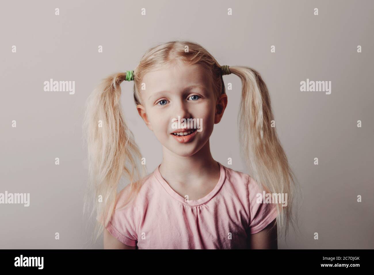 Closeup portrait of surprised sad blonde Caucasian preschool girl in pink t-shirt on light background. Child with long pig tails hair posing Stock Photo