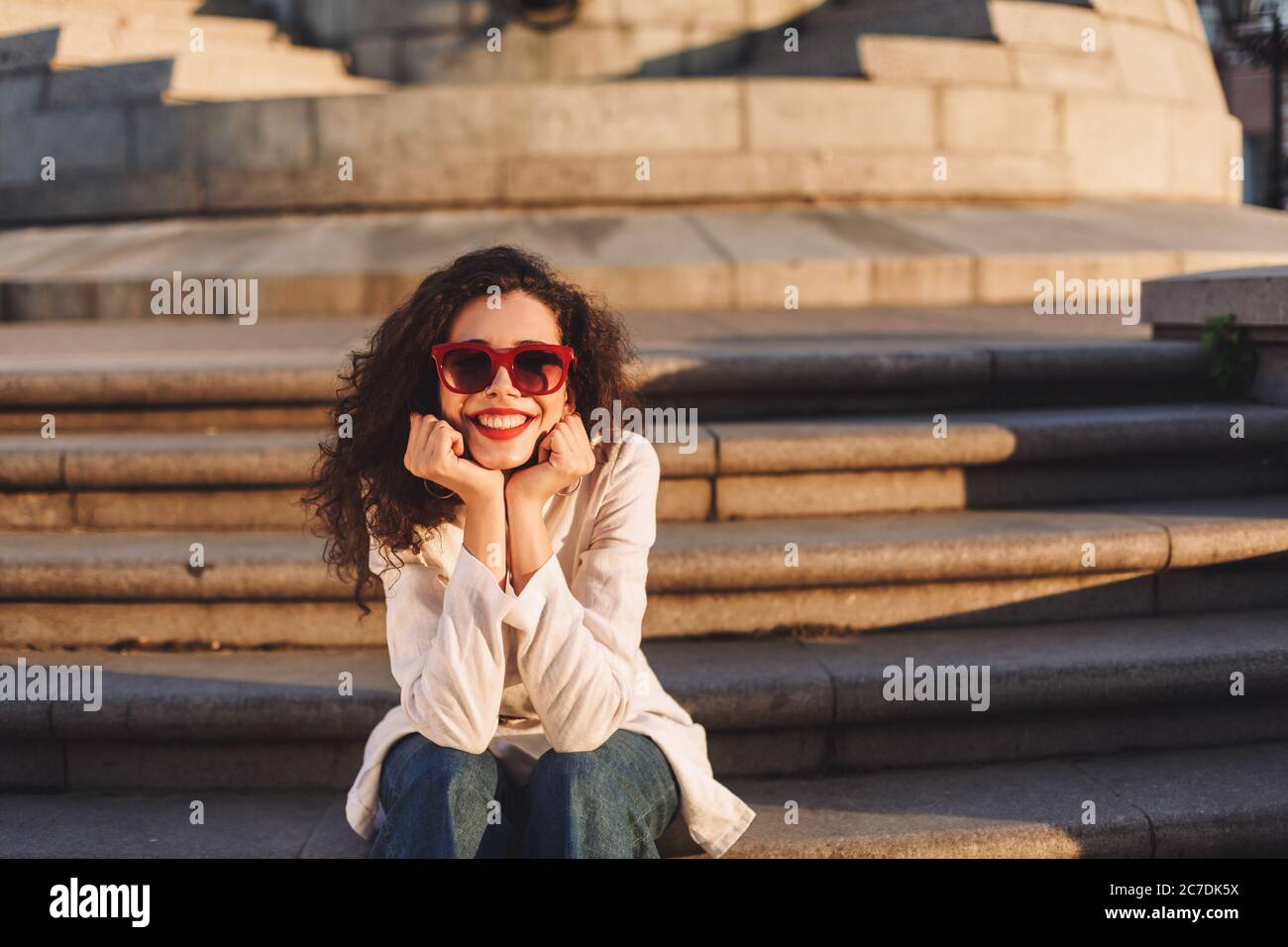 Beautiful lady with dark curly hair in sunglasses and white jacket sitting on stairs with cellphone and listening music in headphones on street Stock Photo