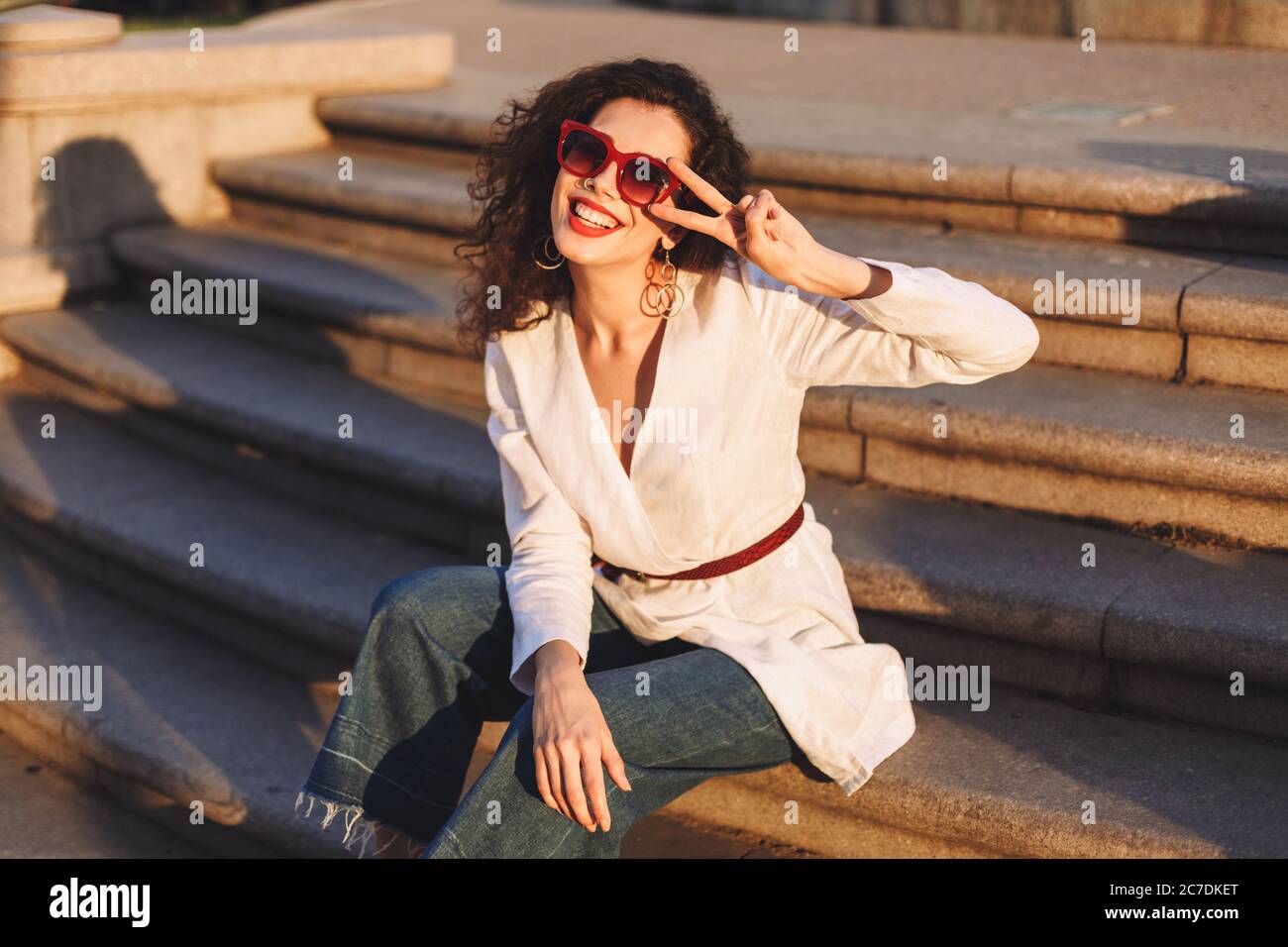 Joyful lady with dark curly hair in sunglasses and white jacket sitting on stairs on street and happily looking in camera while showing two fingers Stock Photo