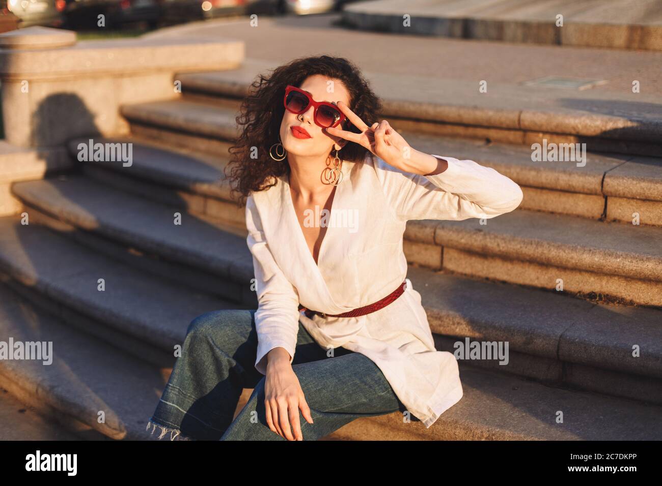 Young beautiful lady with dark curly hair in sunglasses and white jacket sitting on stairs on street and dreamily looking in camera while showing two Stock Photo