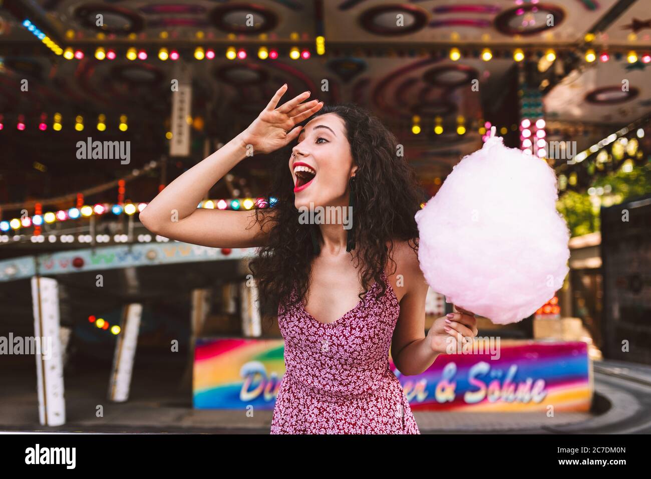 Young joyfull lady with dark curly hair in dress standing with pink cotton candy in hand and happily looking aside while spending time in amusement Stock Photo