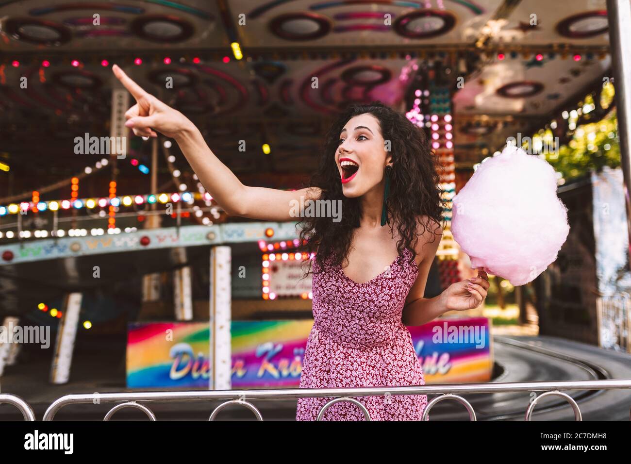 Young joyfull woman with dark curly hair in dress standing with cotton candy in hand and happily showing index finger aside while spending time in Stock Photo