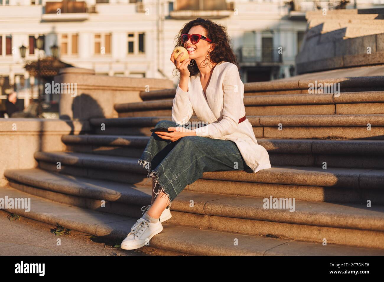 Beautiful smiling lady with dark curly hair in sunglasses and white jacket sitting on stairs with cellphone and apple in hands on street Stock Photo