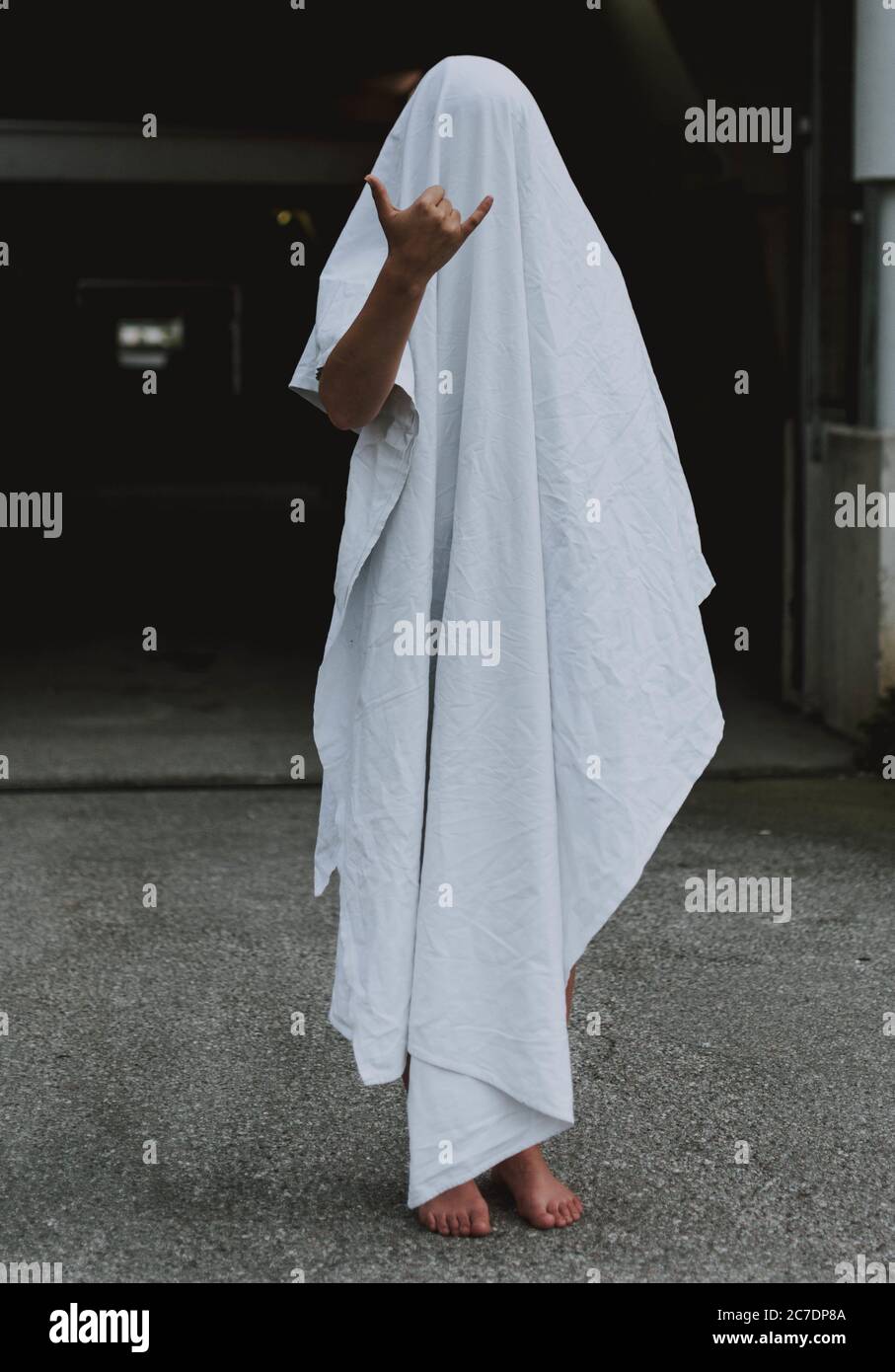 Vertical shot of a barefoot person wearing white ghost sheet showing shaka sign Stock Photo