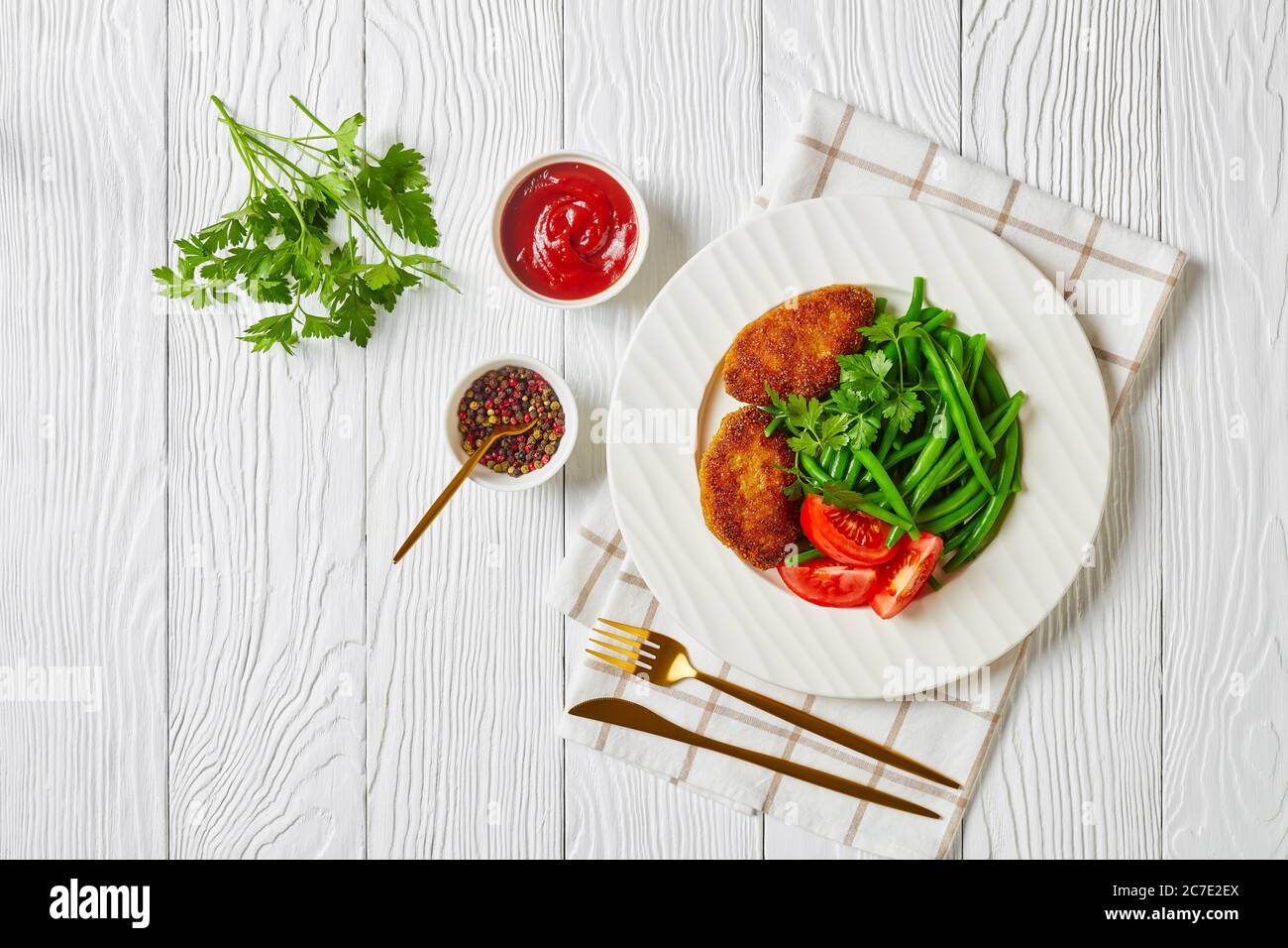 oven baked polenta crusted minced beef patties served with blanched green beans and fresh tomatoes on a white plate on a white wooden table, horizonta Stock Photo
