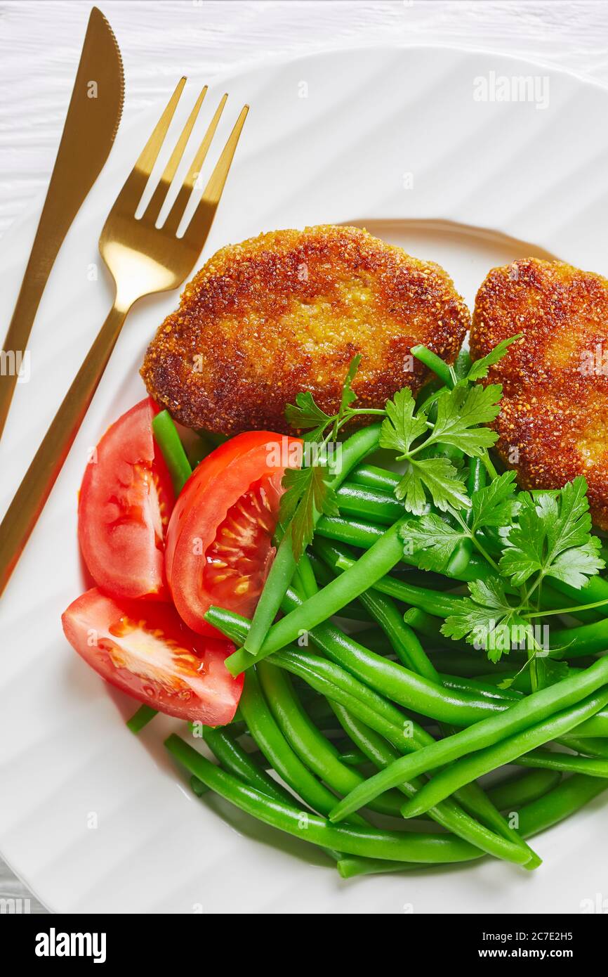 oven-baked polenta crusted minced beef patties served with blanched green beans and fresh tomatoes on a white plate on a white wooden table, vertical Stock Photo