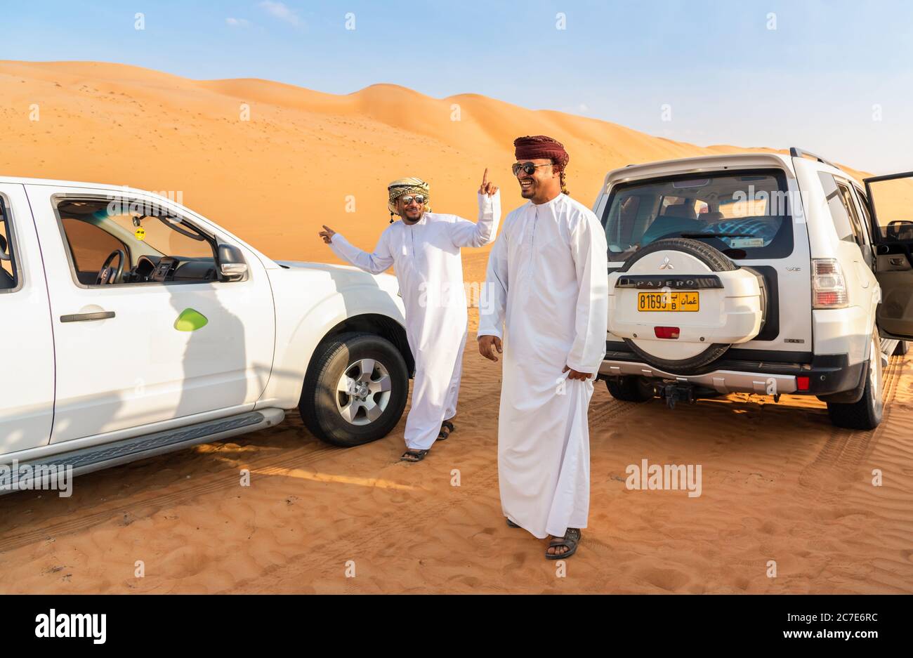 Wahiba Sands, Oman - February 12, 2020: Happy Omani men in traditional clothes have fun with white 4x4 offroad cars in Wahiba Sands Desert, Sultanate Stock Photo