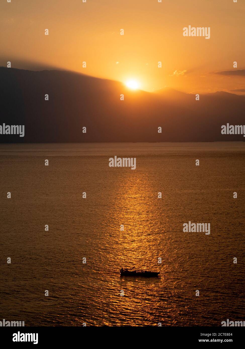 Sun setting behind mountains, reflecting across sea with small boat in the foreground Stock Photo