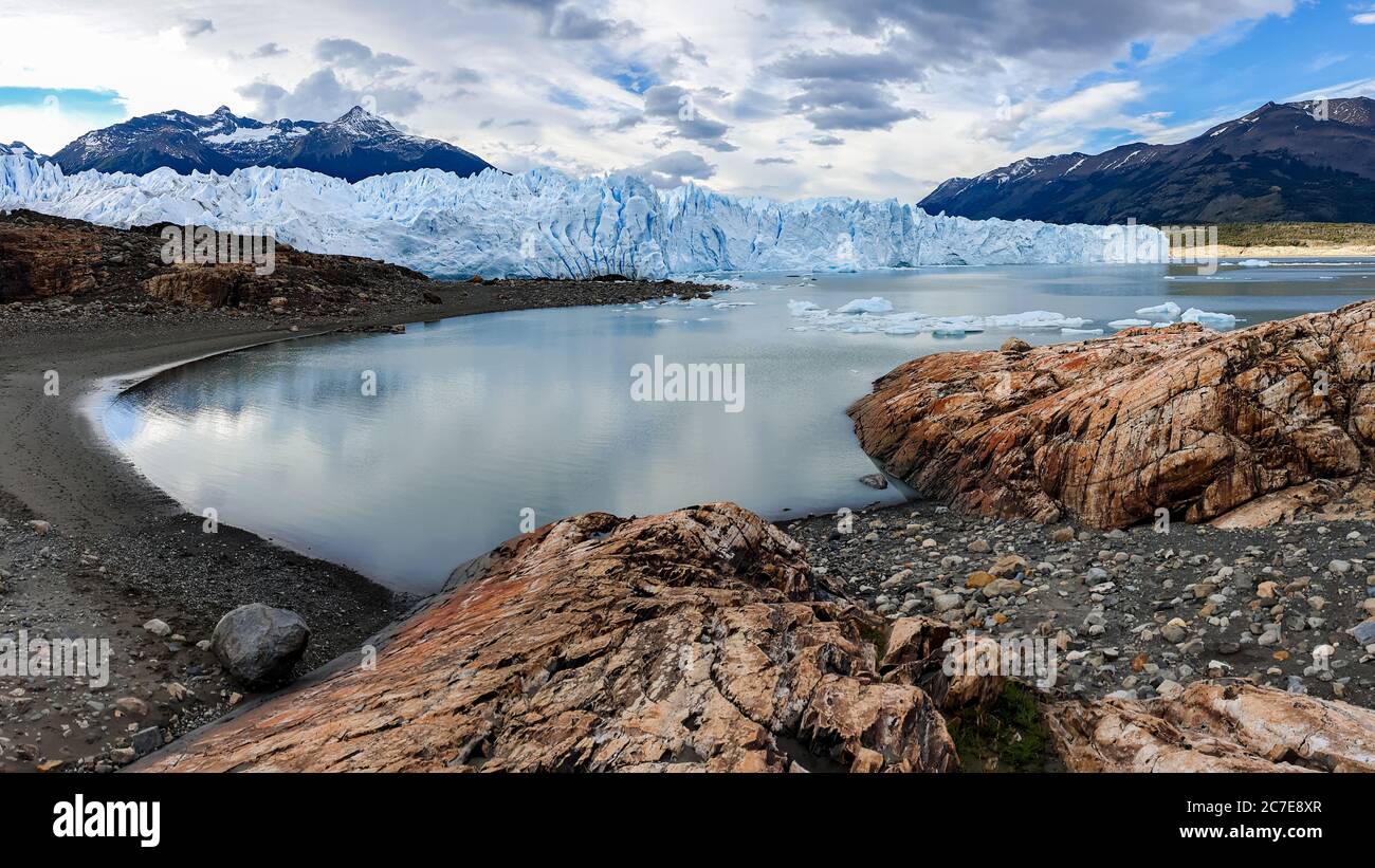 Perito Moreno Glacier reflecting in glacial lake in front of it with rocks in foreground and mountains in the background Stock Photo