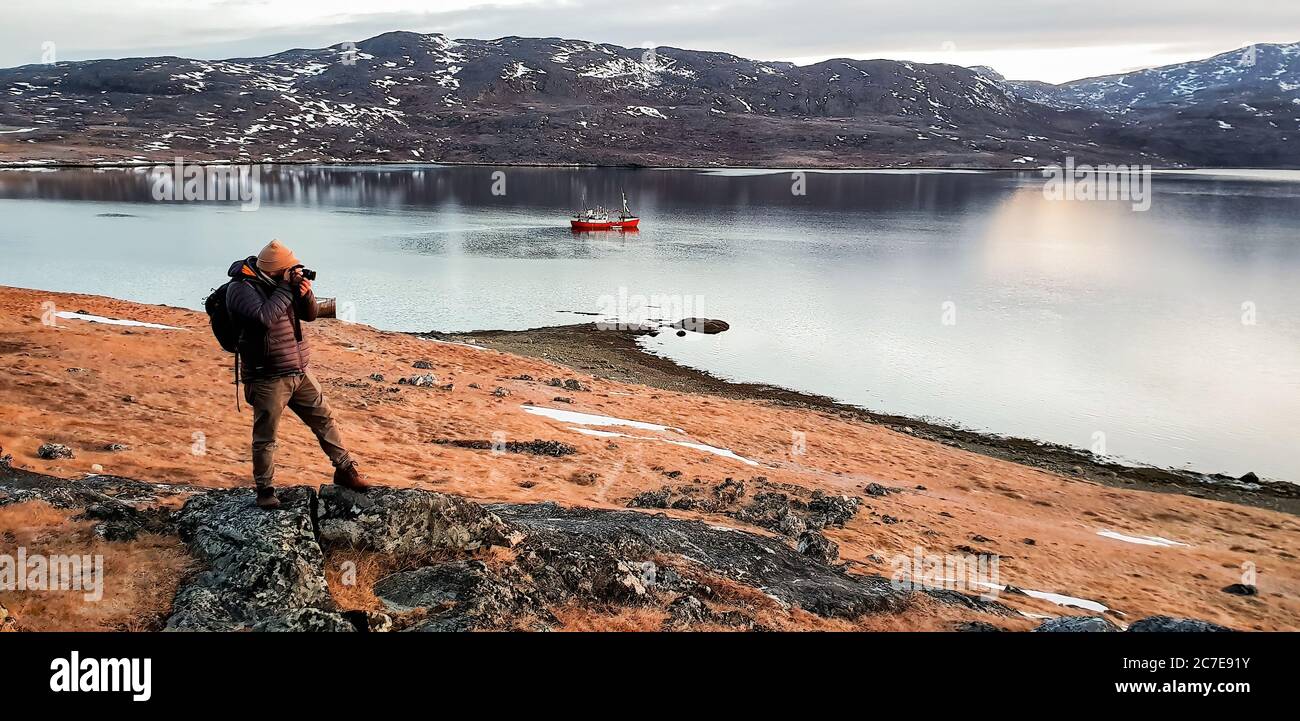 Photographer taking photo whilst stood on rock in Greenland with boat and fjord in the background Stock Photo