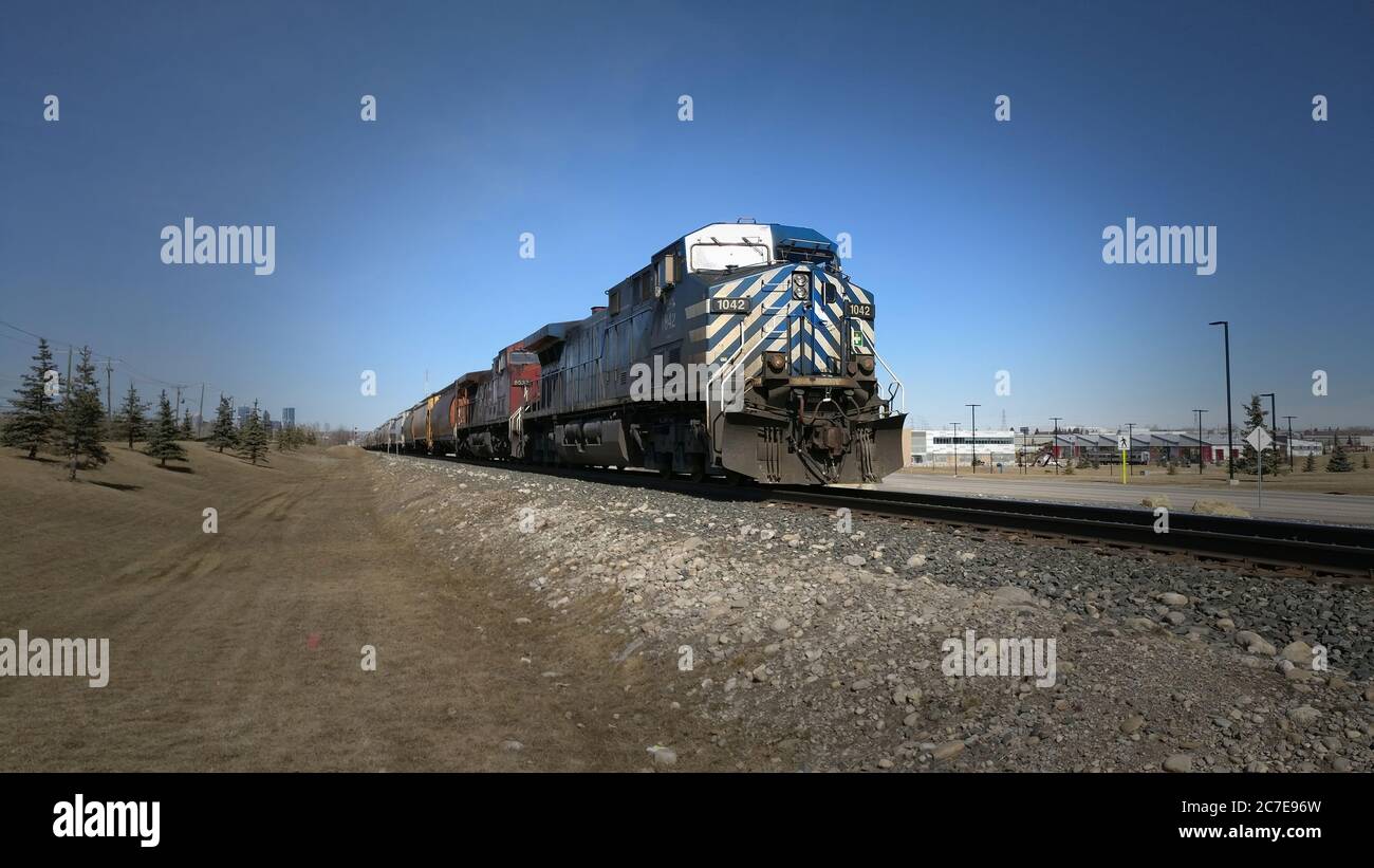 Blue and red train engines pulling mixed cargo cars with buildings in the background Stock Photo