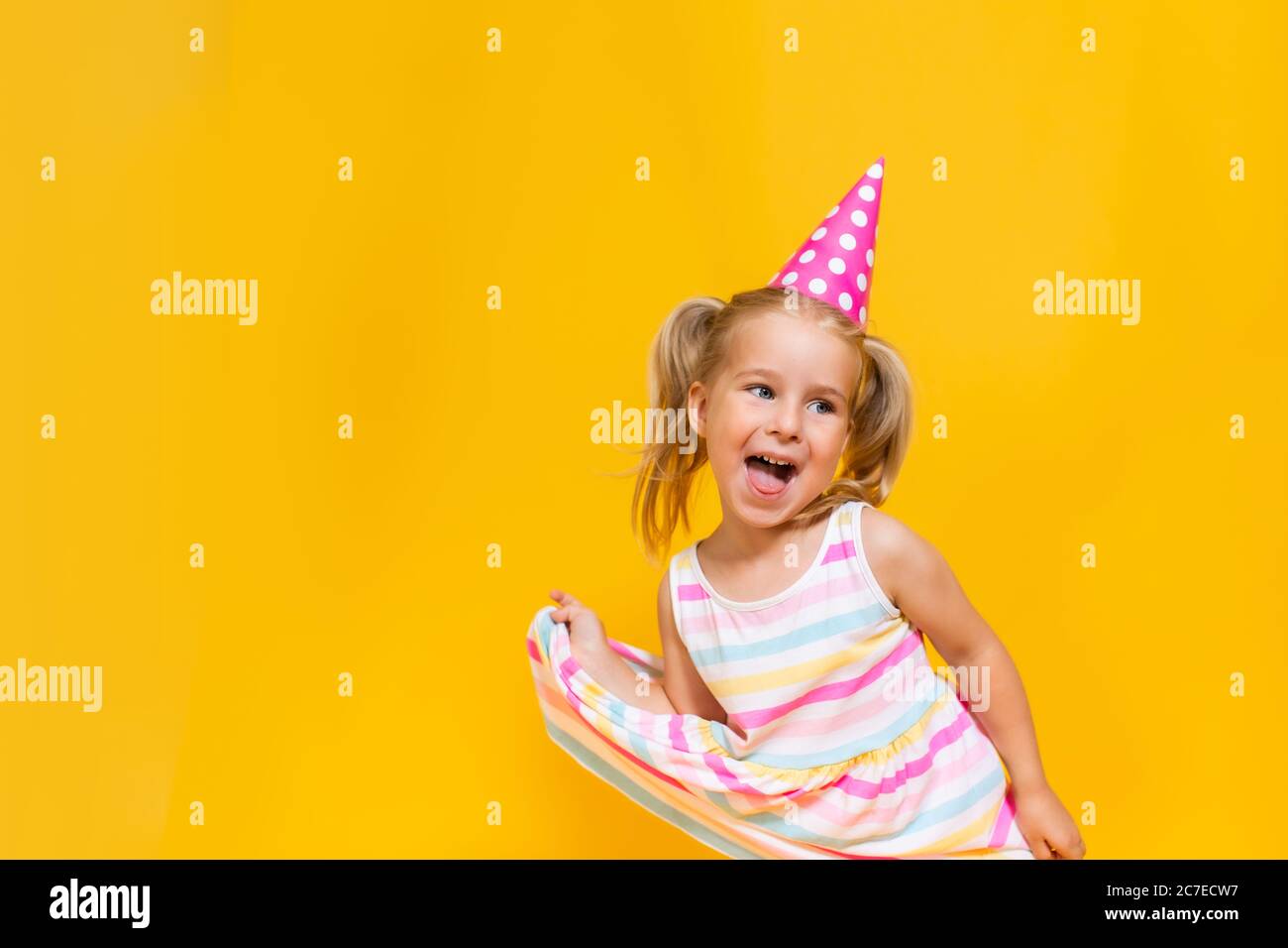 Happy birthday child girl with two ponytales in pink cap on colored yellow background shoing her tongue. Stock Photo