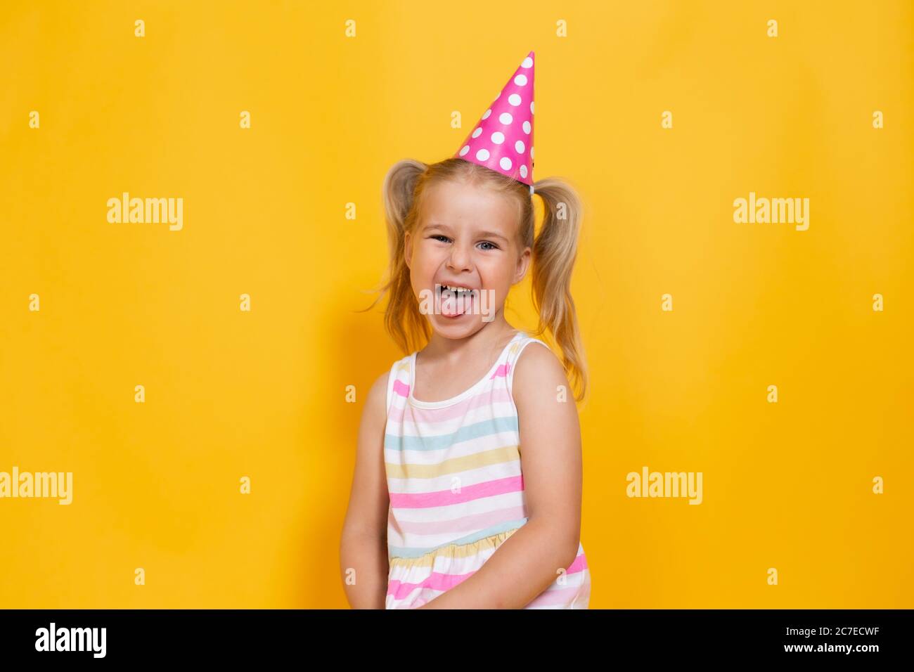 Happy birthday child girl with two ponytales in pink cap on colored yellow background shoing her tongue. Stock Photo