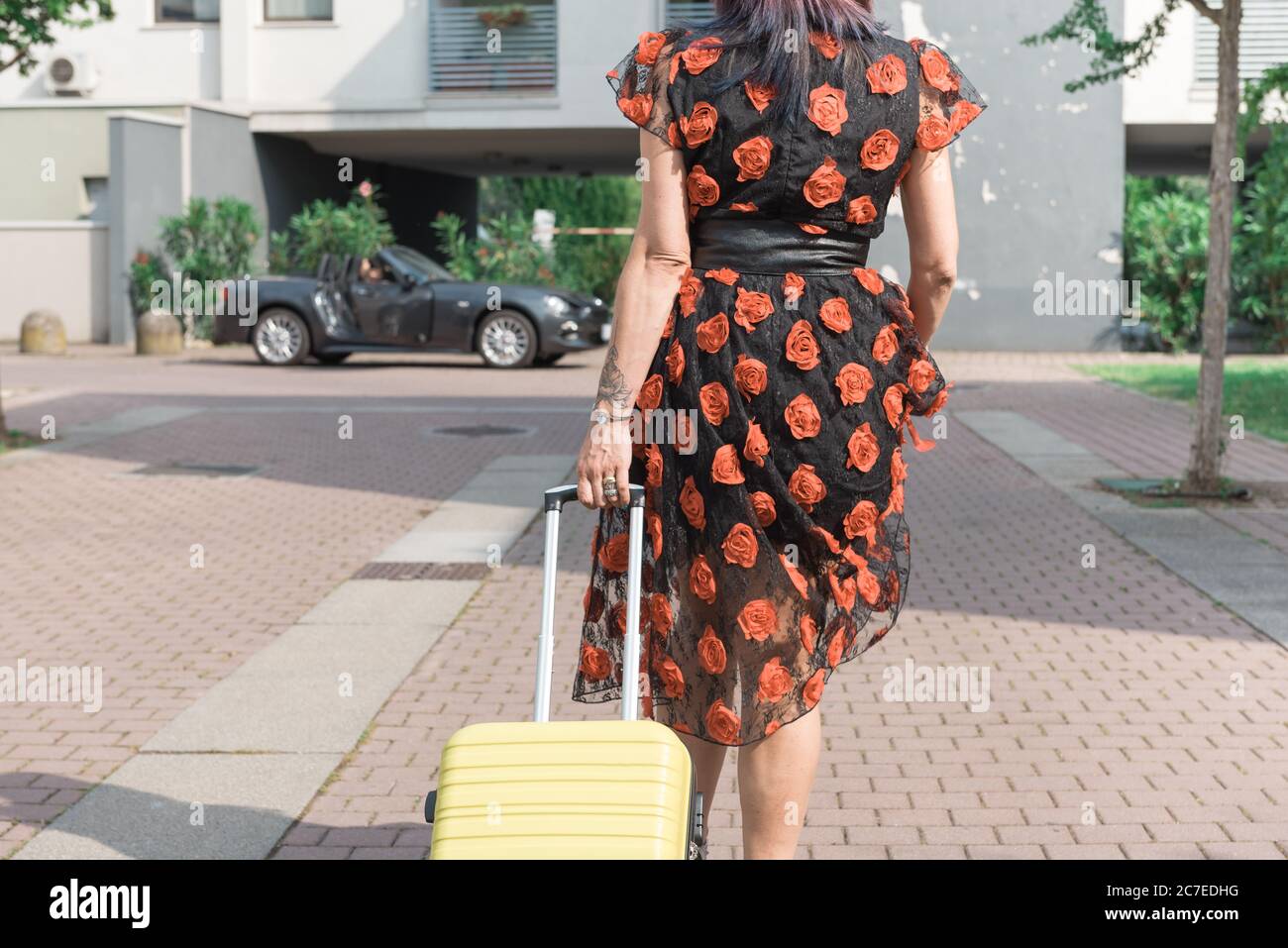 Woman in flowered dress walking towards the car with suitcase. Stock Photo