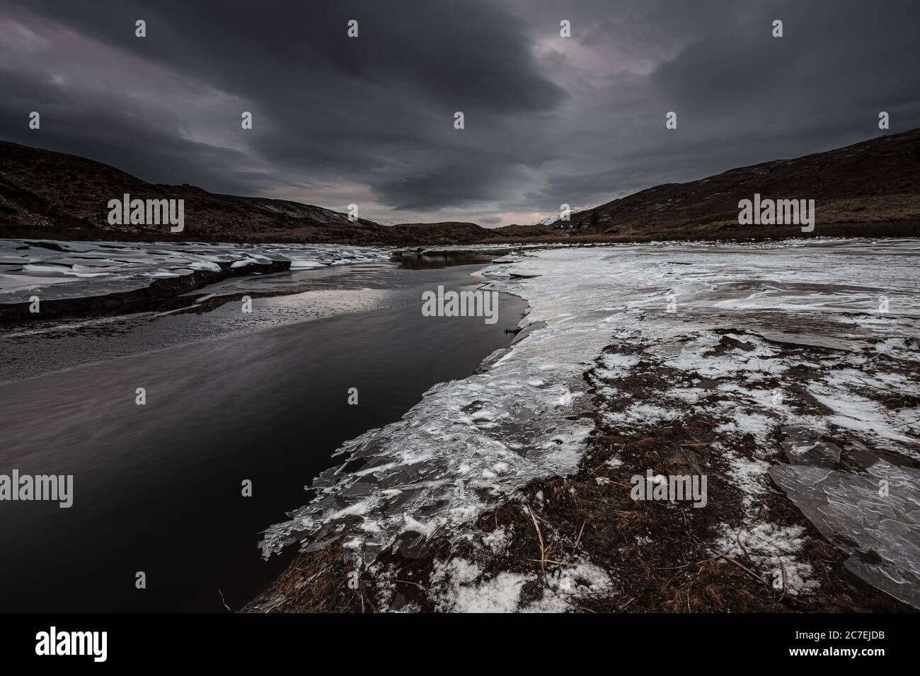 Beautiful shot of frosted surfaces by the water surrounded by mountains under a gray cloudy sky Stock Photo