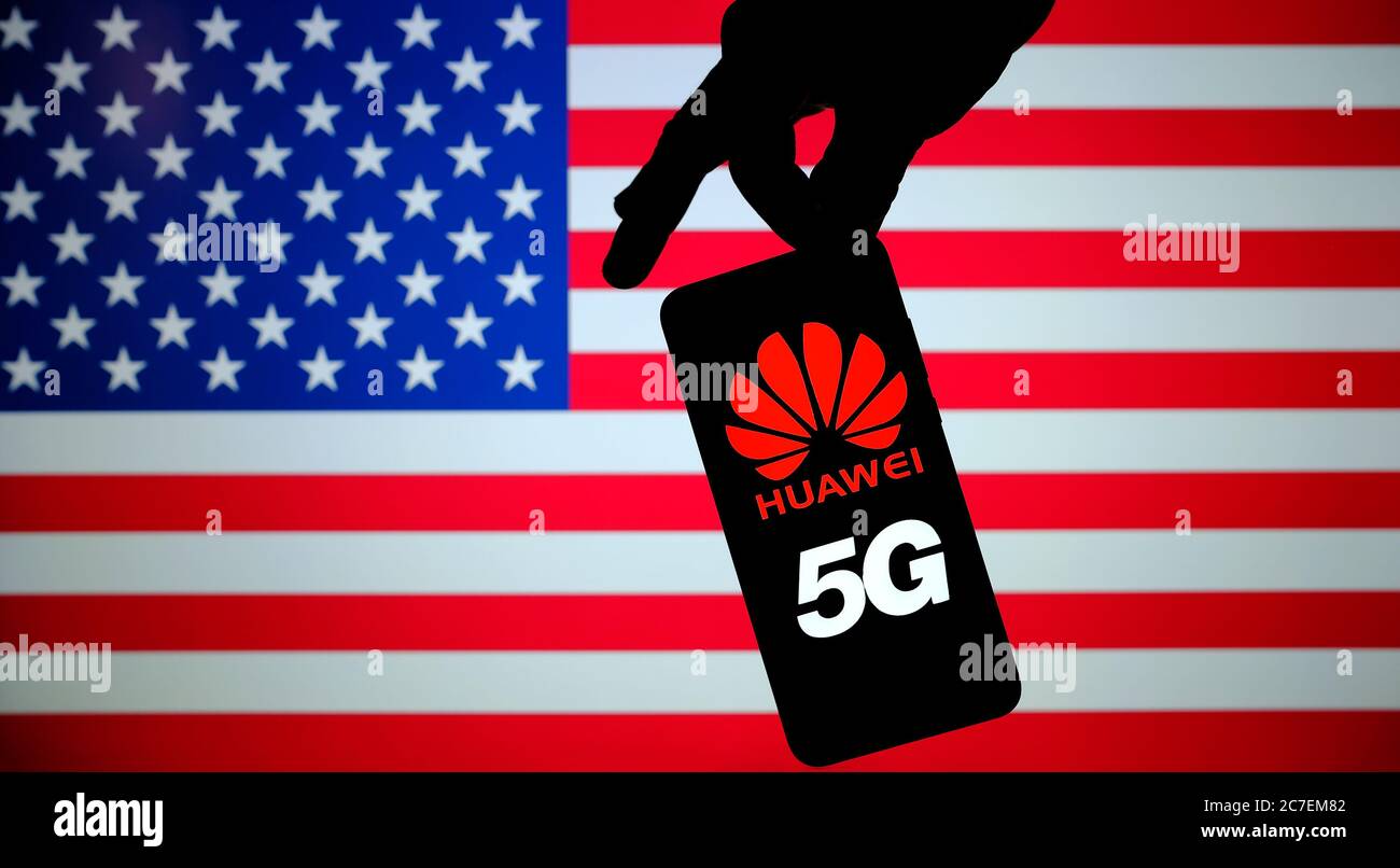 Stone / UK - July 16 2020: Huawei 5G logo seen on smartphone silhourtte hold in hand with the US blurred flag on the background. Stock Photo