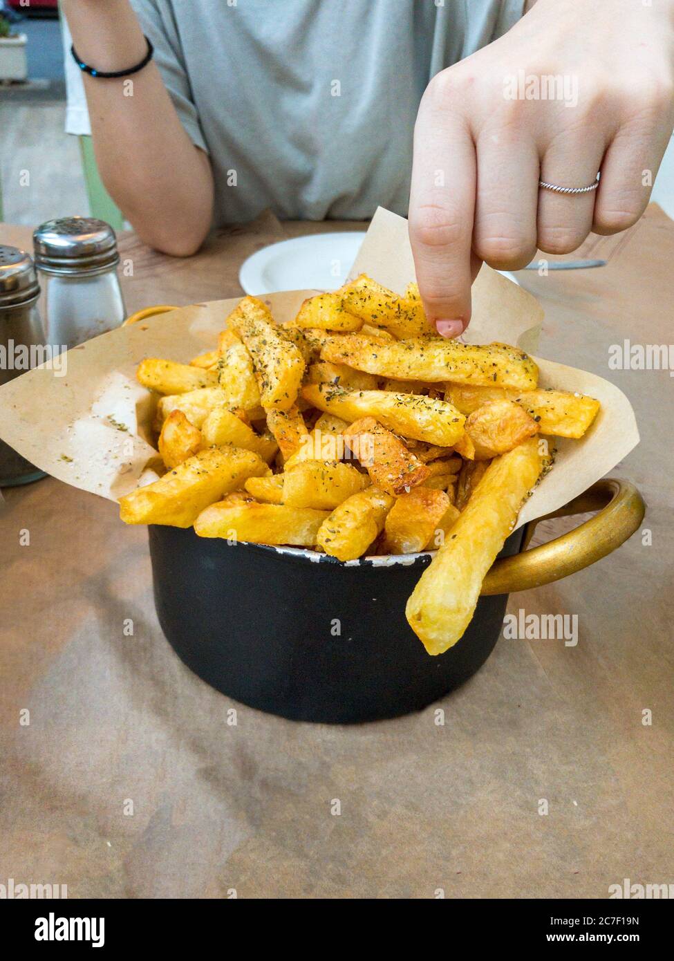 Close-up of a female young woman taking a fried potatos with oregan placed on a pan on a table. Stock Photo