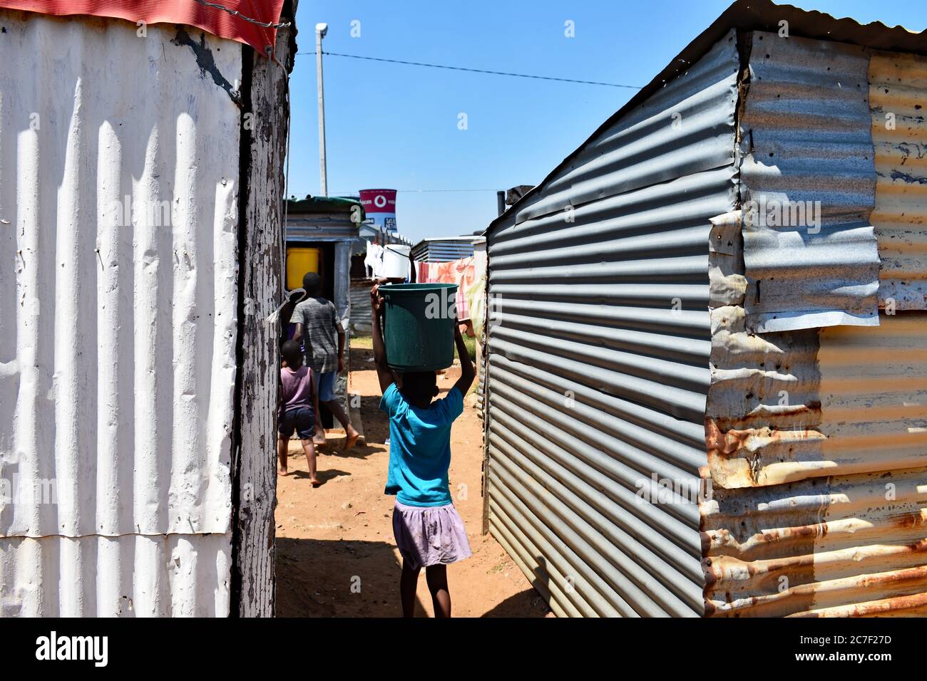 A young girl carries a bucket of water on her head, following her mother and sibling between the slum housing in Soweto, Johannesburg. Stock Photo