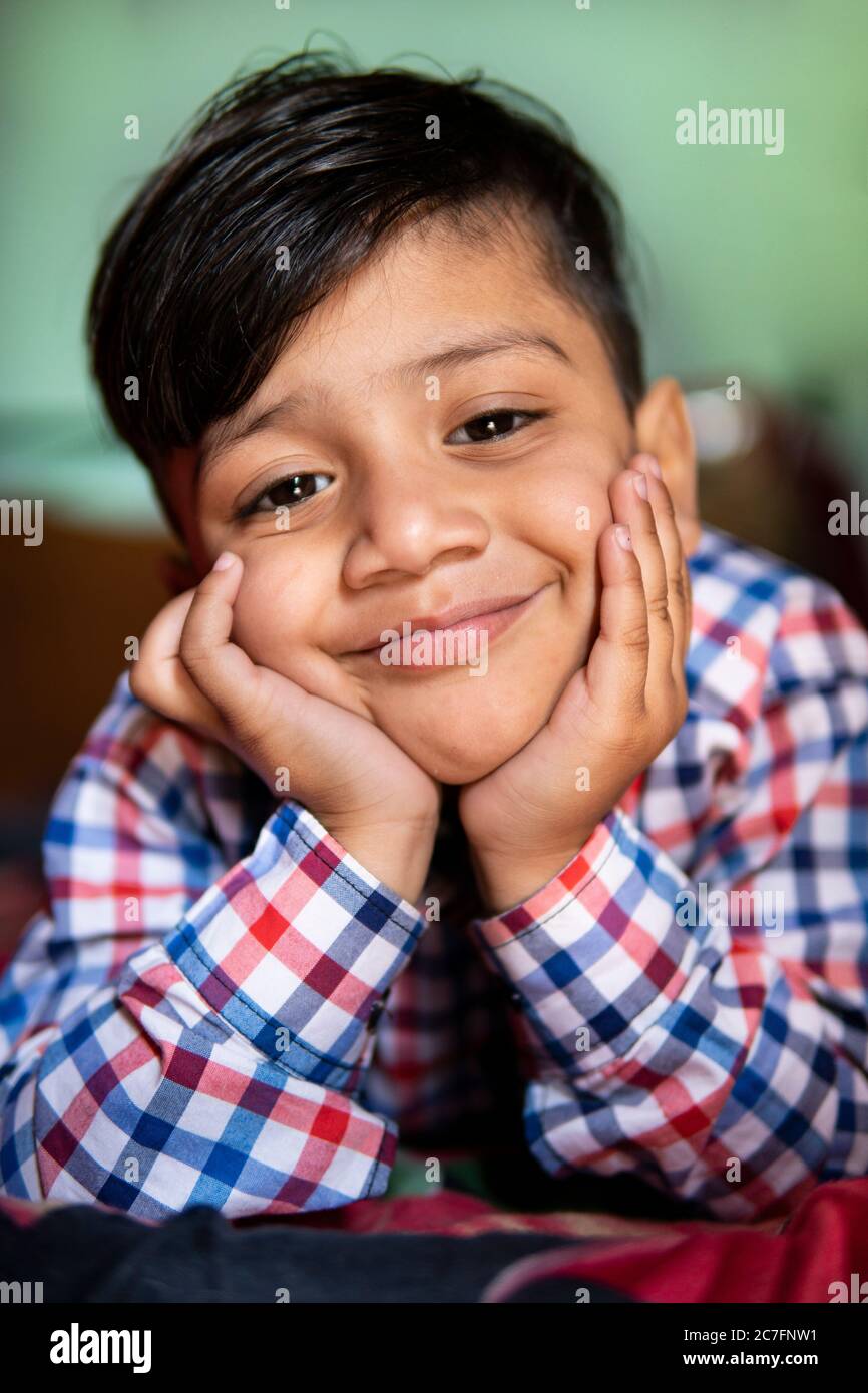 Portrait of cute happy little boy lying front on bed at home. He is relaxing with hands on chin and looking at camera with smile. Stock Photo