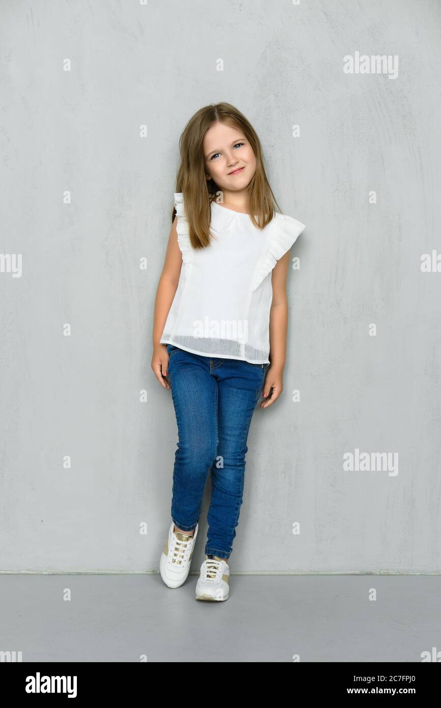 Little girl in white blouse and jeans leaning to the wall in