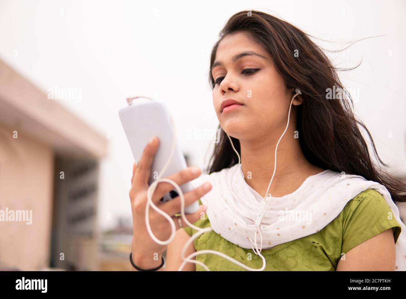 Beautiful, serene Indian late teen girl listening music on smartphone through earphones and enjoying fresh air in outdoor on roof at day time. Stock Photo