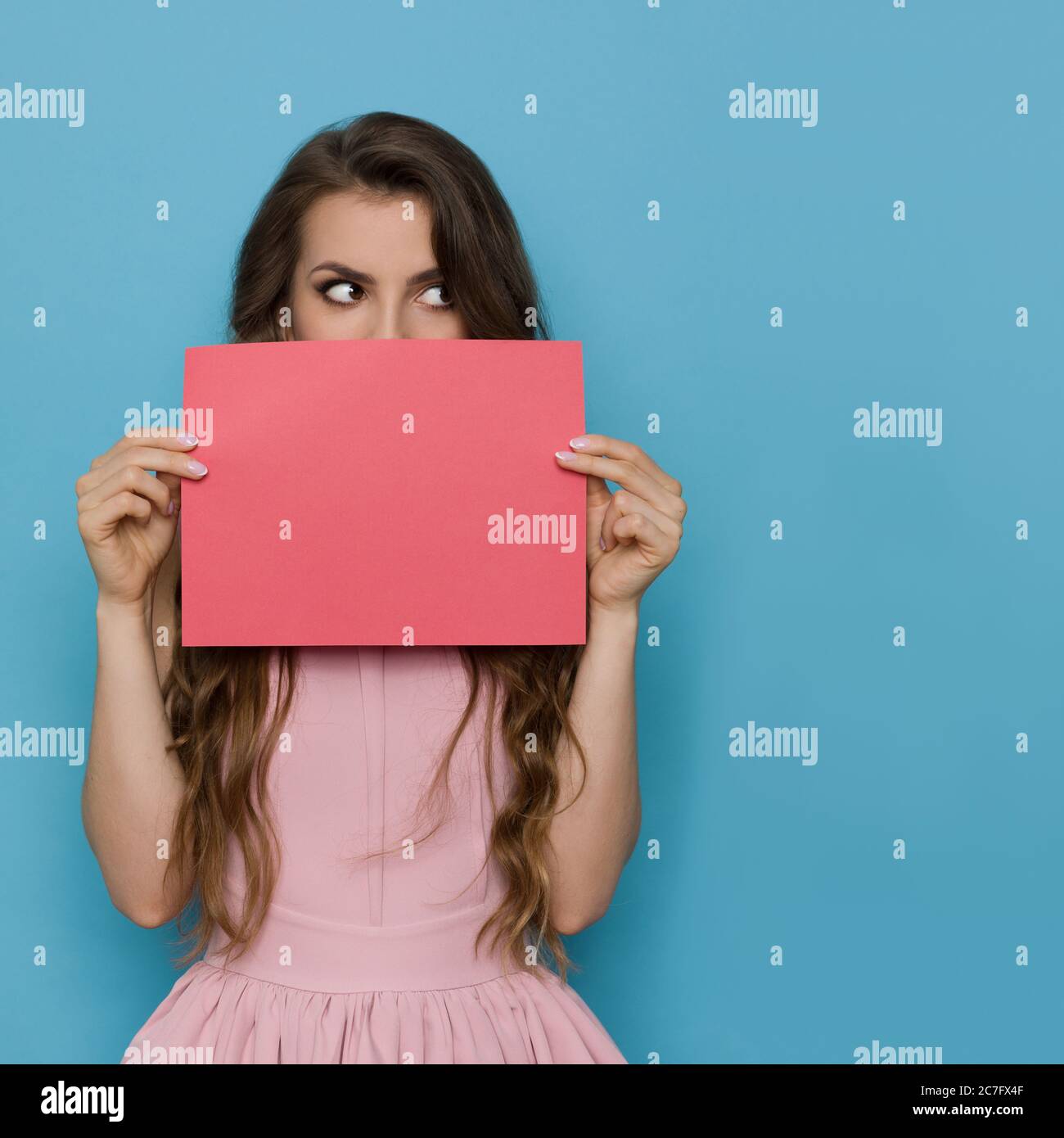 Elegant young woman in pink cocktail dress is hiding her face behind empty sheet of paper and looking at the side. Waist up studio shot on blue backgr Stock Photo