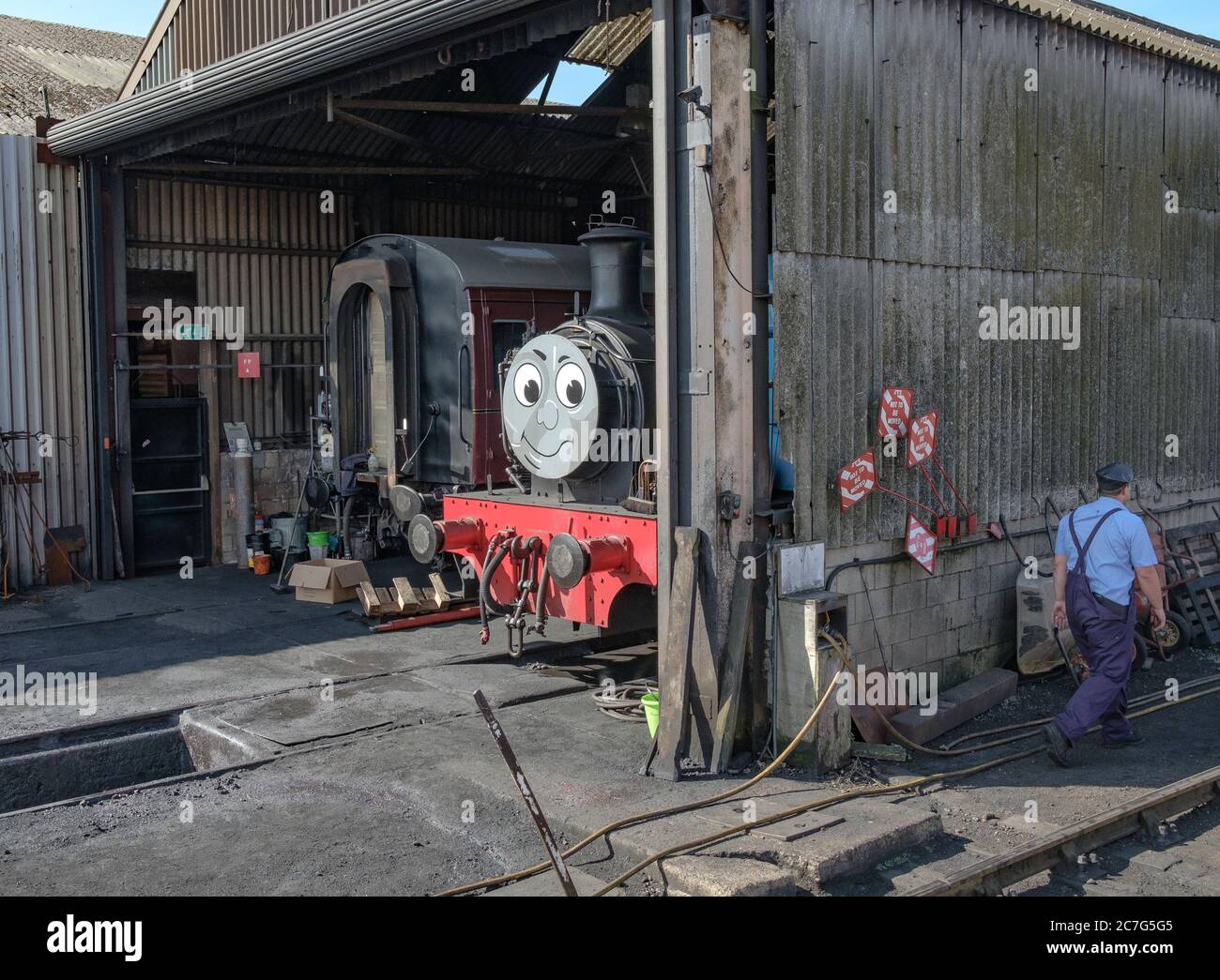 Vintage styled steam locomotives including a popular children's character train seen in a locomotive shed with a train driver seen walking past. Stock Photo
