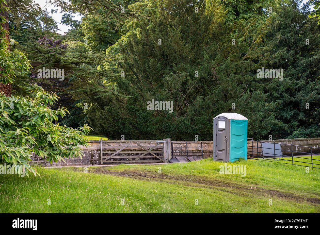 A plastic portable toilet next to a muddy track in a field at an outdoor event and surrounded by tall trees. Stock Photo