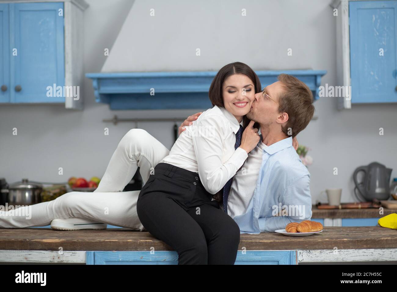 Happy couple higging in kitchen at home. Romantic couple in kitchen sitting on counter. Happy man kissing his gorgeous wife while laying on modern Stock Photo