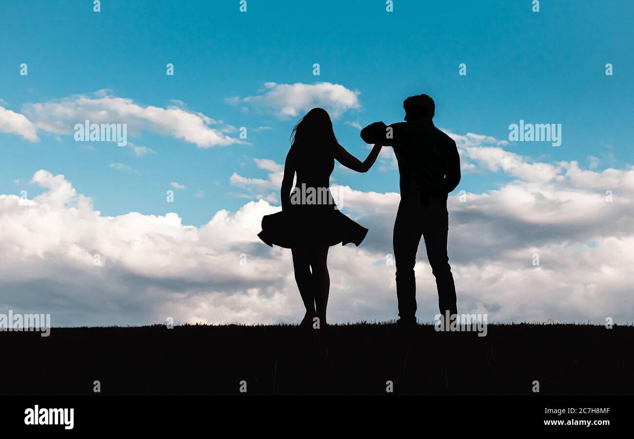 Silhouette of man and woman dancing against a blue sky with clouds. Stock Photo