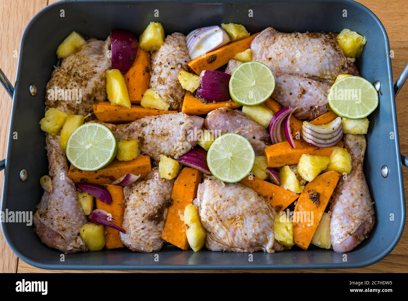Jerk chicken thighs & drumsticks tray bake in roasting tin ready to cook with vegetables: sweet potatoes, red onions, lime & pineapple chunks Stock Photo