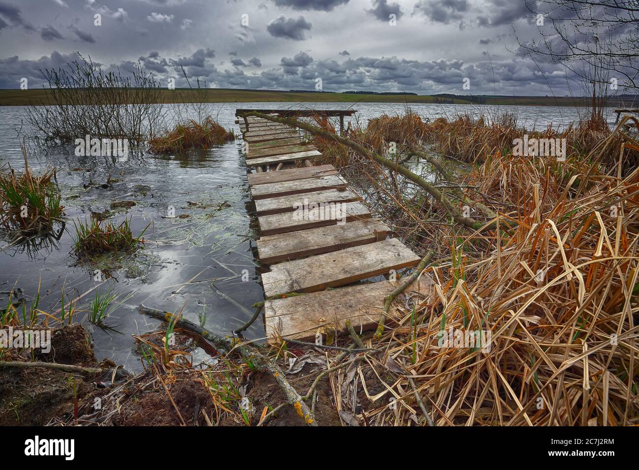 Old wooden pier with dry reed on sunset. Cloudy weather Stock Photo