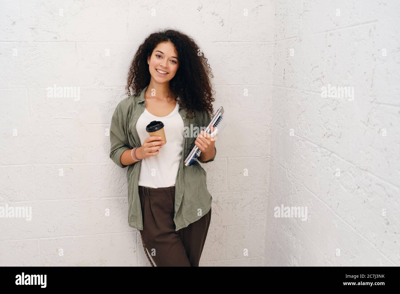 Young pretty smiling woman with dark curly hair in khaki shirt holding notepads and cup of coffee to go in hands happily looking in camera Stock Photo