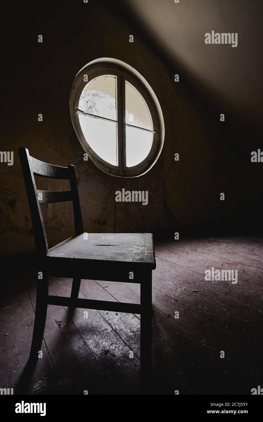 Vertical shot of a wooden chair in a closed area with a round window - depression, solitude concept Stock Photo