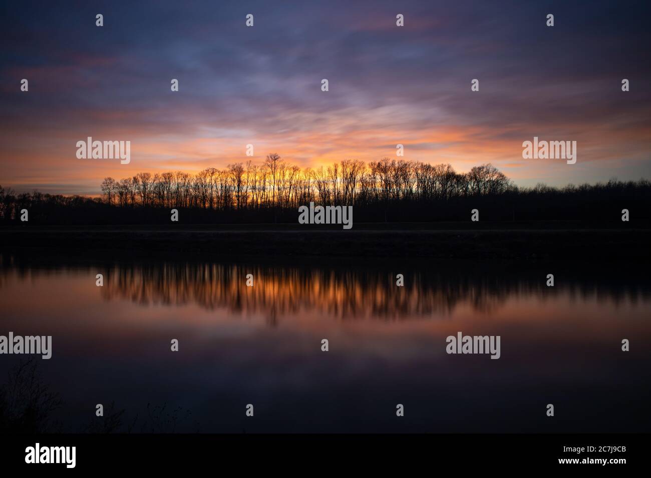 Silhouettes of trees under the cloudy sunset sky reflected in the lake ...