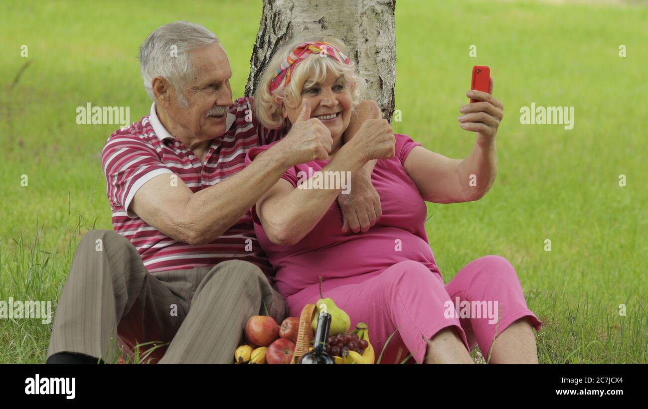 Family weekend picnic. Old people, merry senior caucasian couple using smartphone makes selfie. Remote online video chat call on phone. Elderly man, woman. Husband and wife in park. Grandparents Stock Photo