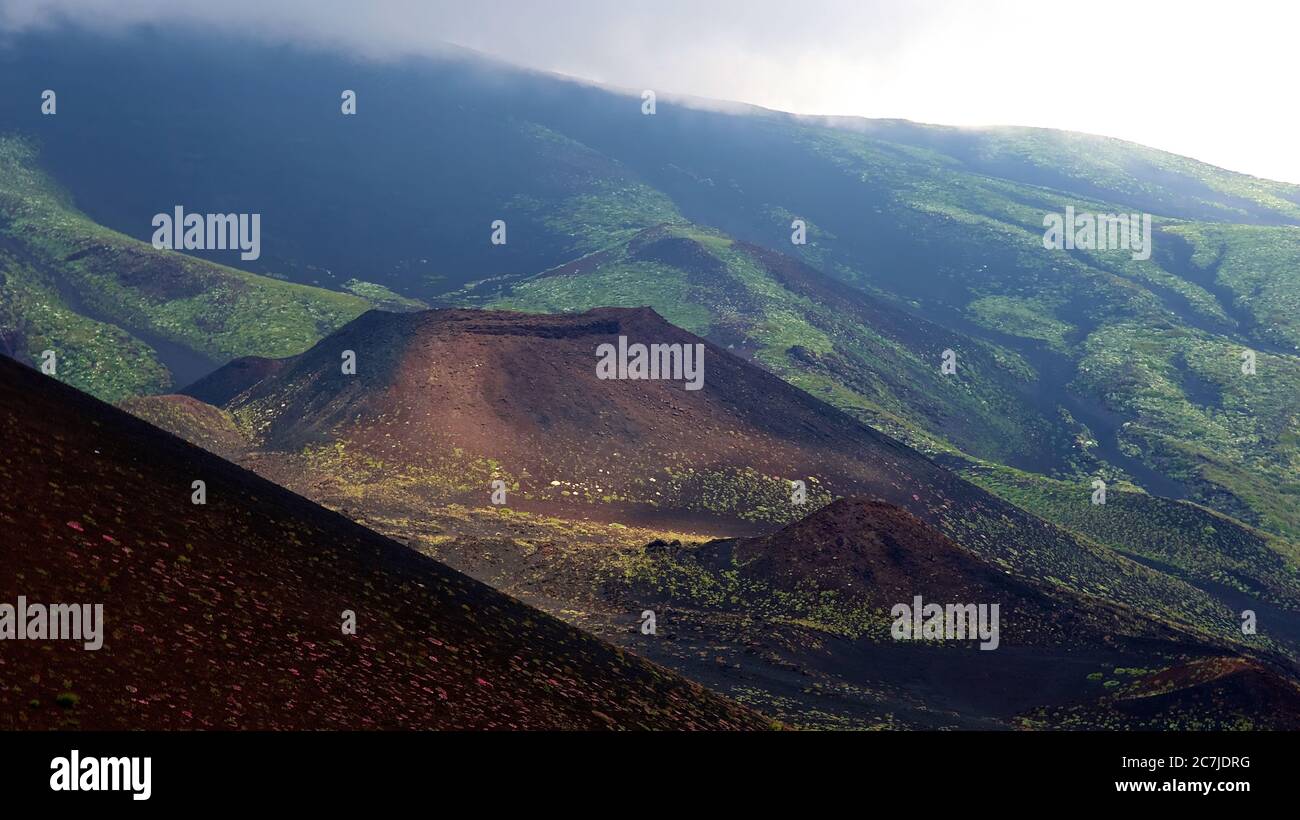 Etna, Etna National Park, Parco dell –´Etna, dark brown lava slope in the foreground, dark, small red crater in the light in the middle, green mountain landscape in the background, cloudy sky Stock Photo