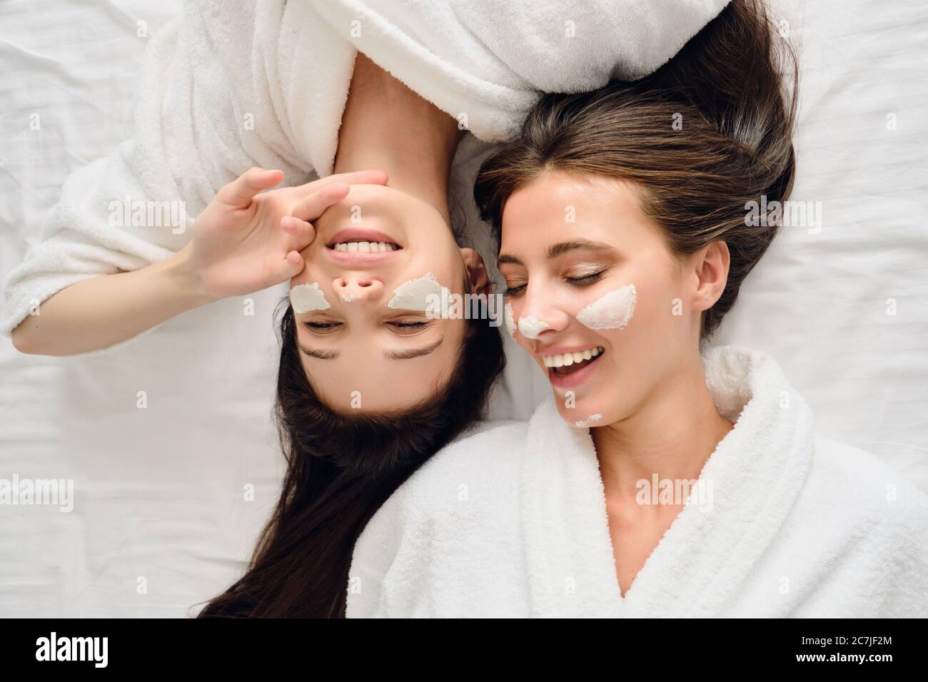 Two young gorgeous smiling women with dark hair in white bathrobes lying in bed with cosmetic mask on faces dreamily closing eyes together in modern Stock Photo
