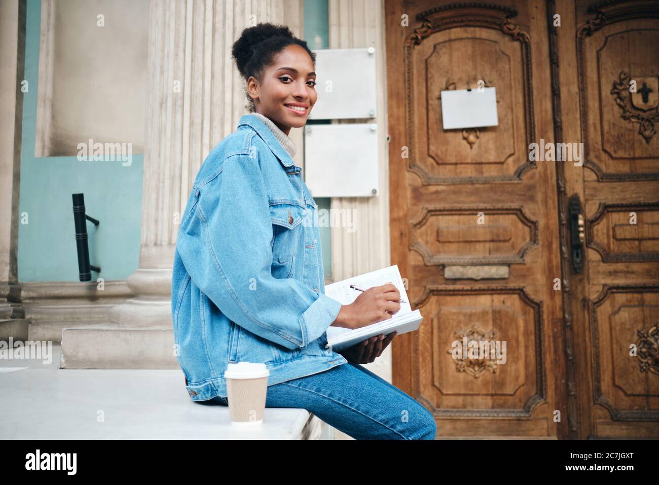 Beautiful smiling casual African American student girl in denim jacket with notebook joyfully looking in camera outdoor Stock Photo