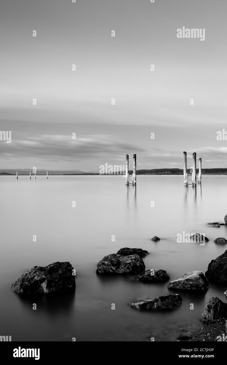 Vertical greyscale shot of a wooden pier near rock formations under the cloudy sky Stock Photo