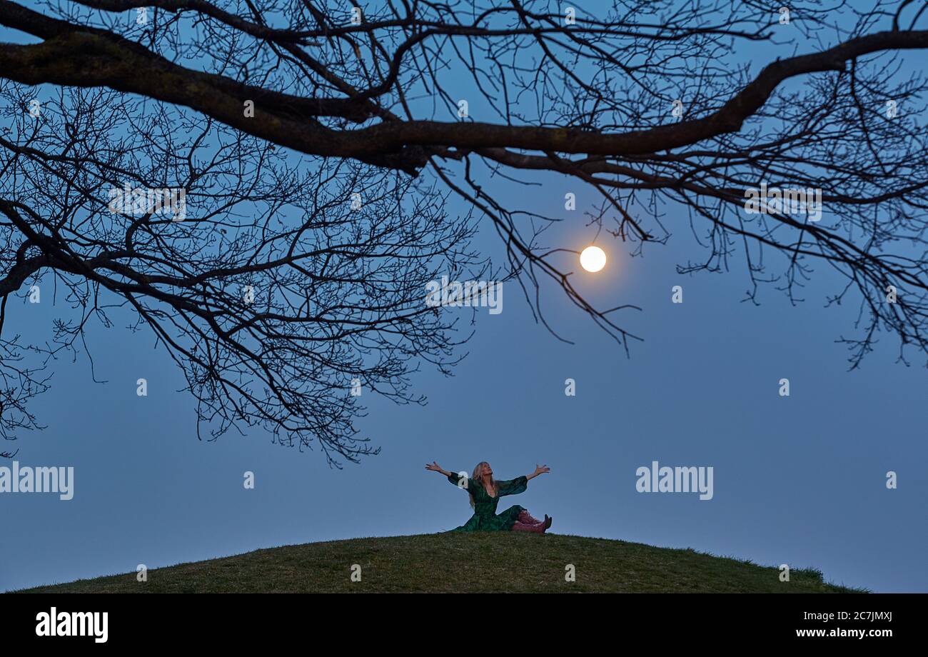 Full moon, branch, trees, glowing, hill, woman, sitting, arms outstretched, looking up at the sky Stock Photo
