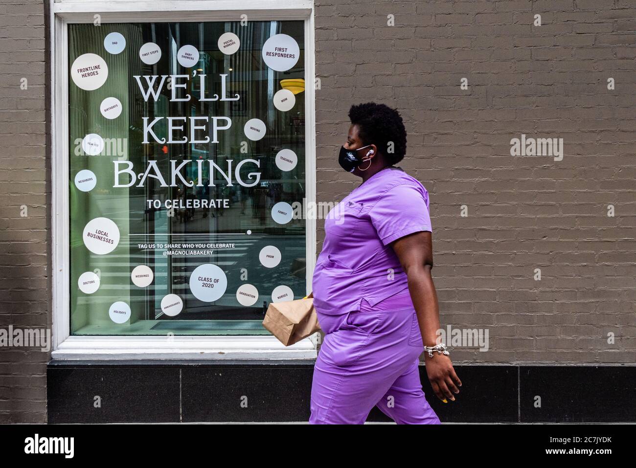 A message in the window of Magnolia Bakery informs customers that they keep on baking despite COVID-19 while a nurse wearing a face mask walks past the store on July 17, 2020, in New York City. (Photo by Gabriele Holtermann/Sipa USA) Credit: Sipa USA/Alamy Live News Stock Photo
