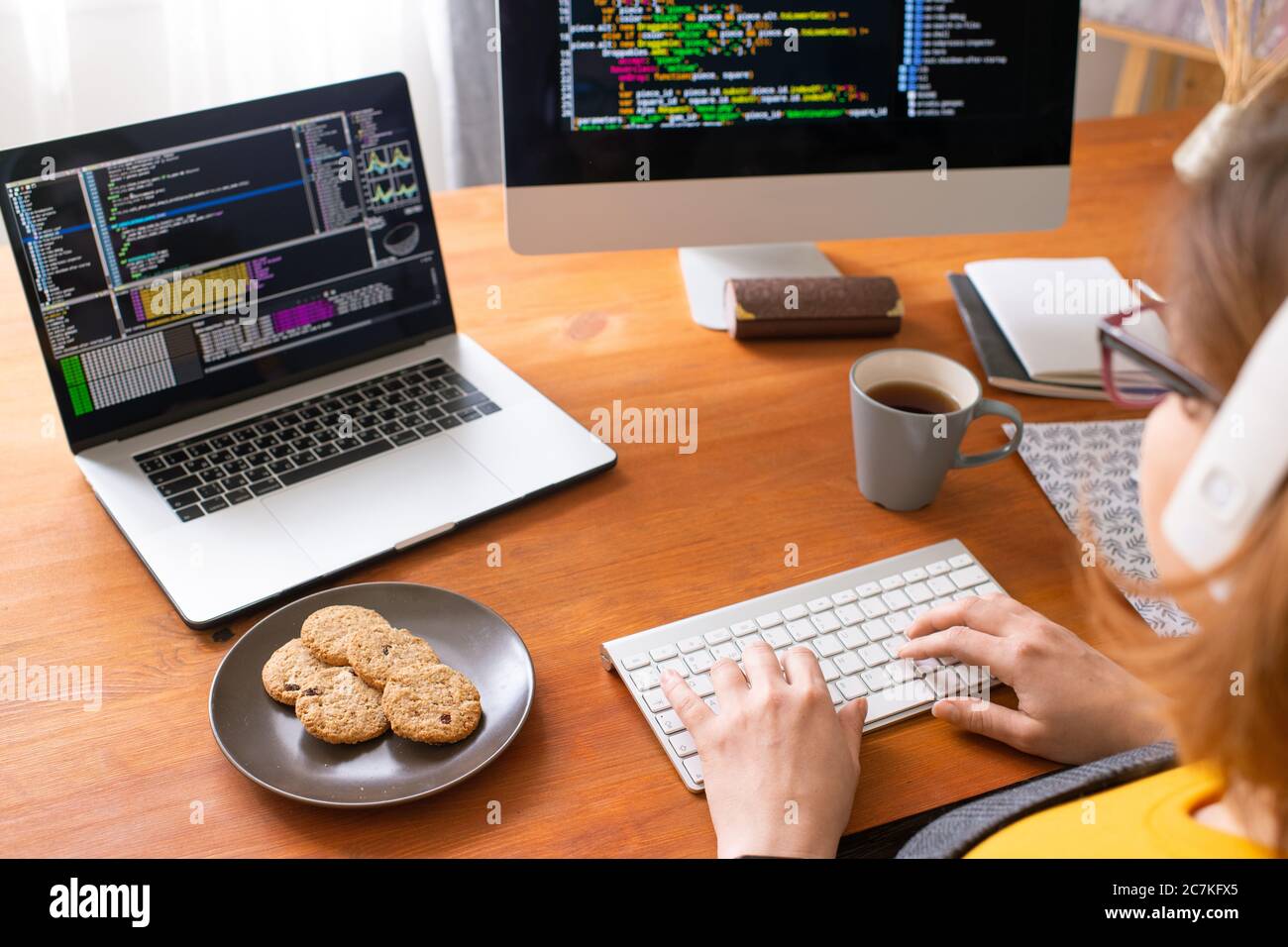 Young female programmer sitting by workplace at home, having tea with cookies and typing on computer keypad while analyzing data Stock Photo